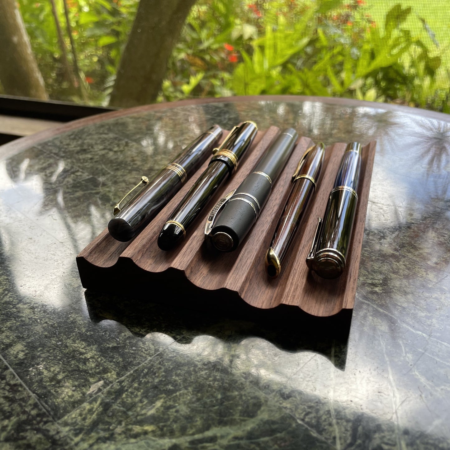 Five black and gold fountain pens arranged on a scalloped wooden display stand, placed on a dark reflective tabletop with a blurred outdoor garden background.