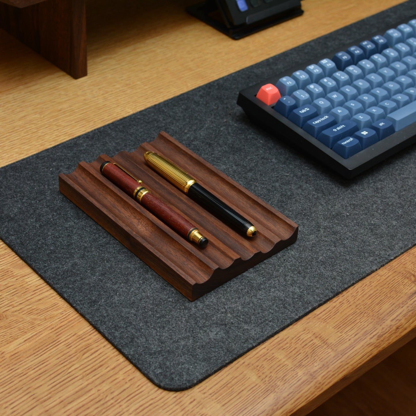 A dark wood pen stand holding two pens on a gray desk mat over a wooden desk, with part of a blue mechanical keyboard visible.