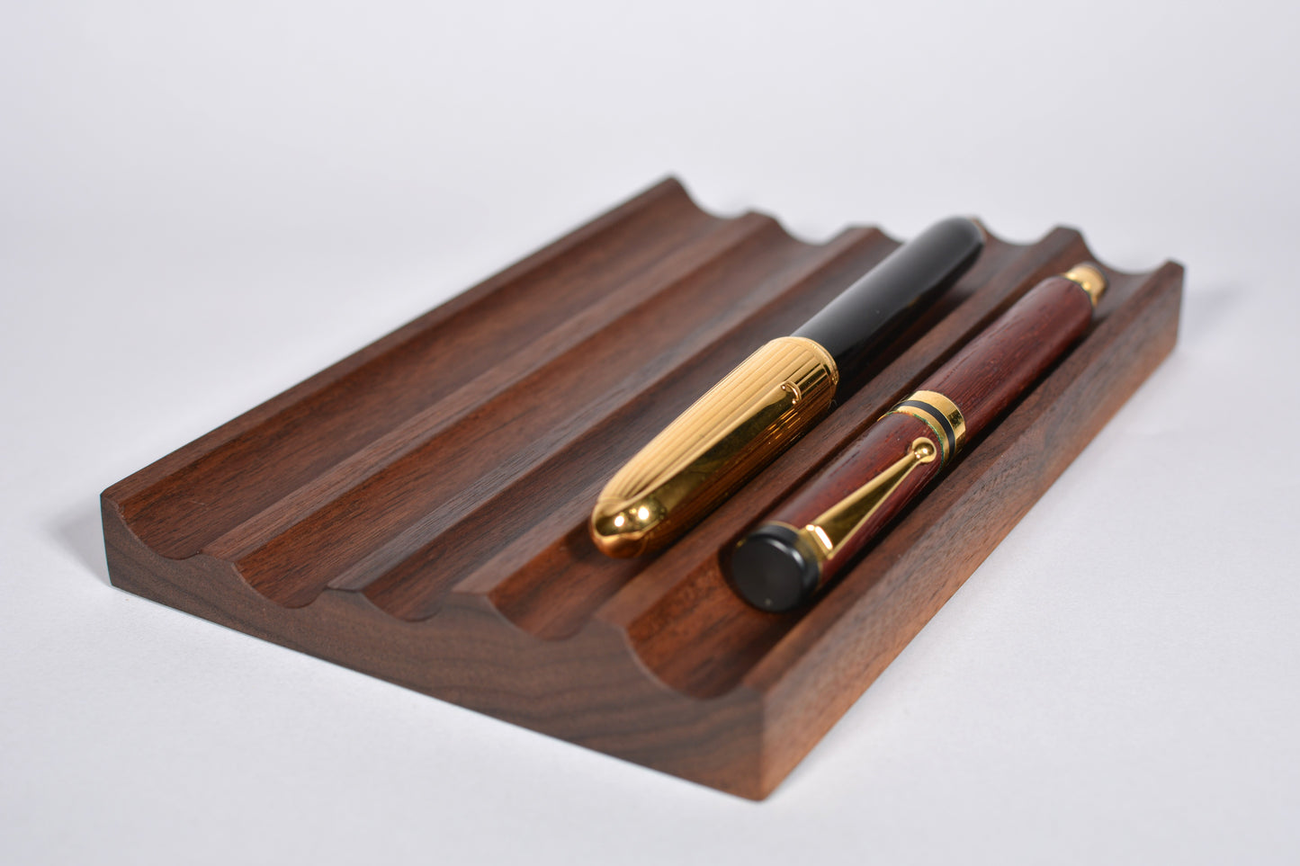 A dark brown wooden pen stand with two elegant pens resting on it, photographed on a clean light gray background.