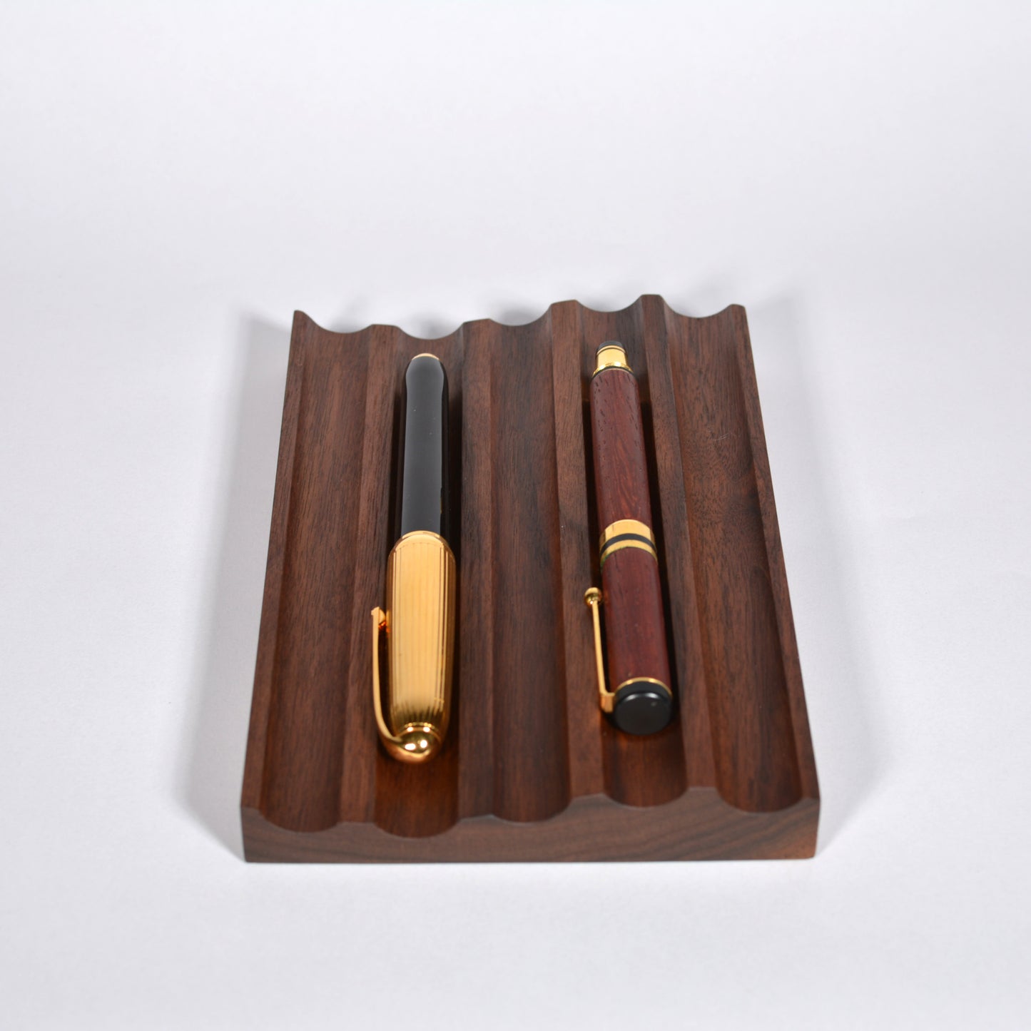 A dark brown wooden pen stand with a wavy slotted design, holding two elegant pens, one with a black and gold barrel and the other with a walnut wood grain and gold trim, set against a plain light gray background.